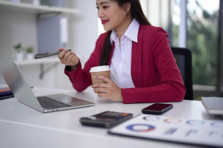 Adult Asian professional woman working on laptop in modern office holding pen and coffee cup for presentation and strategic planningの写真素材