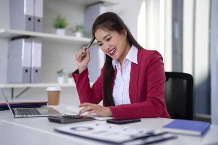 Adult Asian businesswoman smiling calculates budget analyzing financial reports with pen notebook laptop modern corporate workspaceの写真素材