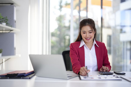 Smiling Asian adult woman works with business reports and calculator in modern office setting. Focuses on financial analysis and strategic planning. Bright workspace.の写真素材