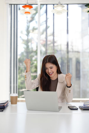 Adult Asian woman expresses joy excitement achieving success working remotely on laptop. Her professional triumph brightens the modern workspace.の写真素材