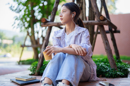 Thoughtful Asian young woman enjoying outdoor park campus environment. Relaxing with coffee listening music near laptop and notebook. Student lifestyle concept.の写真素材