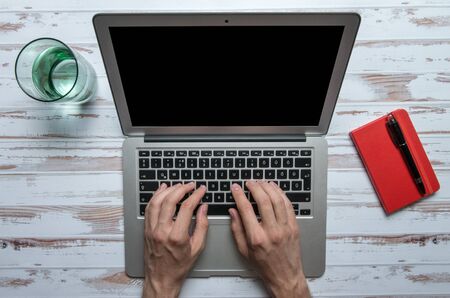 Man typing on a laptop on a white table with a glass of water and a red notebook with a penの写真素材