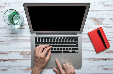Man using a touchpad of a laptop on a white table with a glass of water and a red notebook with a penの写真素材