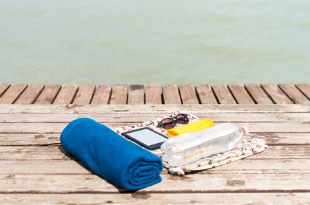 Summer essentials: sunglasses, towel and something to read on a pier with a lake in the backgroundの写真素材