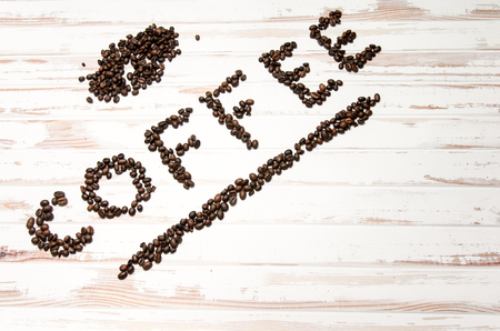 The word coffee written with fresh coffee beans with some extra coffee on a white wooden tableの写真素材