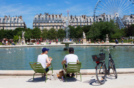 PARIS - JULY, 2016: Man and woman have lunch by pool at Jardin Des Luxembourg. A bike, ferris wheel, fountain and buildings by the park are in the view in Paris.のeditorial素材
