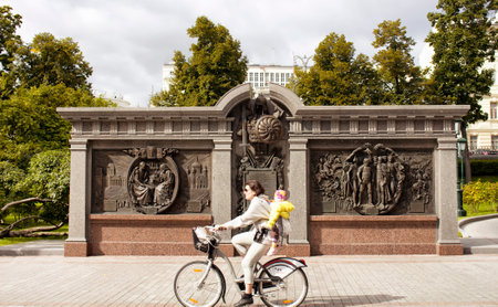 Woman riding bicycle with her daughter passes by monument to Alexander 1 at Alexandrovsky Garden in Moscow. Park with lawns, summer blooms & several memorials including one to Marshal Georgy Zhukov.のeditorial素材