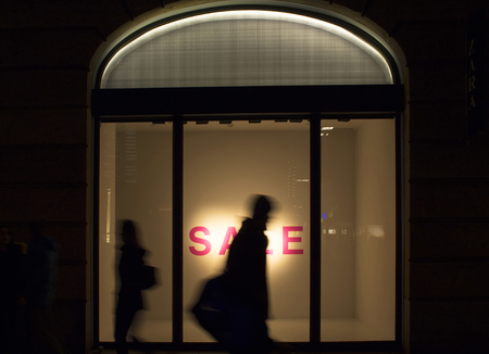 Silhouettes of man and woman walking in blurry motion in front of a fashion store's window in night. Conceptual image of sale season, consumerism and capitalism of modern world.の写真素材