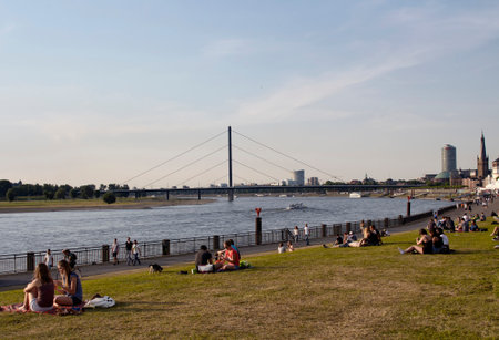People enjoy sunny weather and hang out by Rhine (Rhein) river in Dusselforf. Oberkasseler Brucke bridge is in the background.のeditorial素材