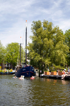 View of three children swimming in canal, two women taking a sun bath, a boat, trees in Amsterdam. It is a sunny summer day.のeditorial素材