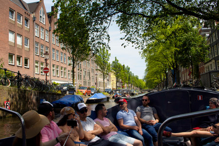 View of people on canal cruise boat tour in Amsterdam. Historical, traditional and typical buildings and trees are in the background. It is a sunny summer day.のeditorial素材