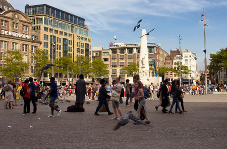 View of people walking at Dam square, pigeons flying in blurry motion. National monument is in the background. It is a sunny summer day.のeditorial素材