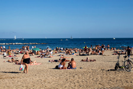 View of many people laying down on sand, taking sunbath and old woman walking at famous beach called "La Barceloneta" in Barcelona. It is a sunny summer dayのeditorial素材