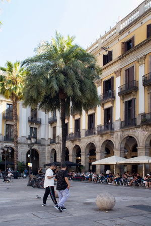View of people hanging out at a famous city square called "Placa Reial", palm trees and historical, traditional buildings in Barcelona. It is a sunny summer day.のeditorial素材