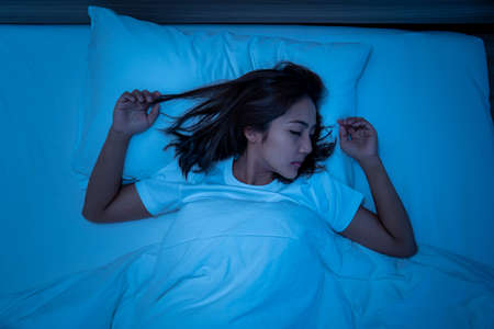 Top View of Beautiful Young Woman Sleeping Cozily on a Bed in His Bedroom at Night. Blue Nightly Colors with Cold Weak Lamppost Light Shining Through the Window.の写真素材