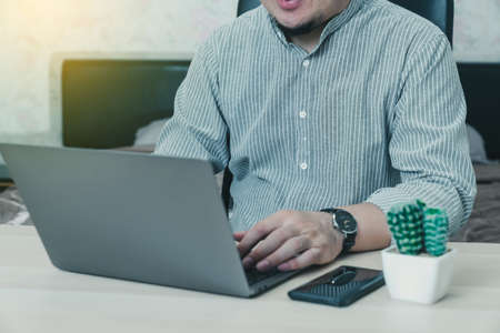 Young businessman in eyewear  working at home with laptop and phone on desk. Pleasant happy man communicating in social network, searching information online.の写真素材
