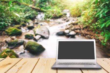 Laptop with blank screen on wooden table with waterfall in forest background, can used for display or montage your productsの写真素材