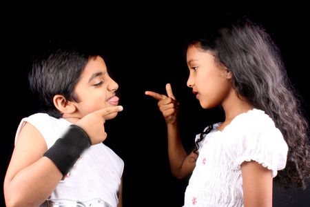 A portrait of two stylish Indian kids posing back to back making faces, on a black background.