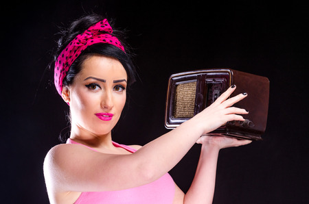 Pin-up style young woman holding a vintage radio against a black backgroundの写真素材