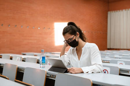 Latin Female Student wearing mask reading a notebook seated on an university bench while pandemicの写真素材