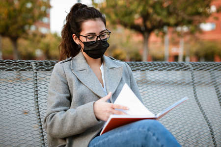Latin Female Student wearing mask reading a notebook seated on an university bench while pandemic.の写真素材