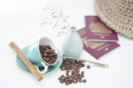 A tabletop scene with light blue coffee cup with coffee beans and cinnamon stick, two British passports and a summer straw hat and a small vase with some wild grass in の写真素材