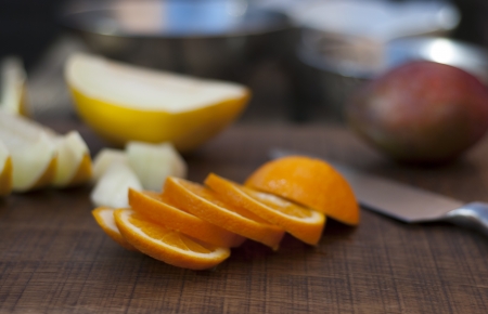 Slices of a orange in a relaxed stack on a chopping board with kitchen knife and soft focused sliced and diced melon, mango and bokeh metal containers in the backgroundの写真素材