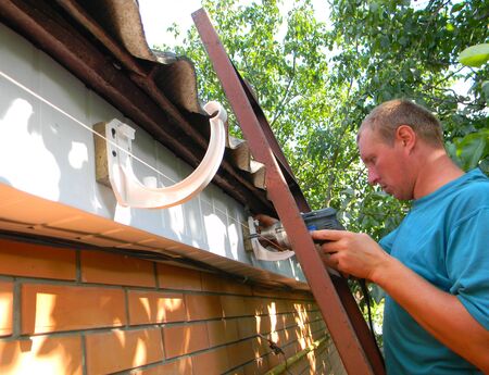 KYIV, UKRAINE - April, 27, 2019: Contractor installing plastic roof gutter holder for downspout...