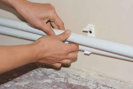 A plumber is installing central heating by laying new white polypropylene pipes into plastic waterline half clamps to create good piping support along the walls in the house.の写真素材