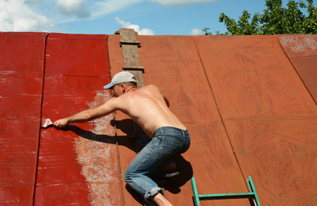 KYIV, UKRAINE - MAY, 24, 2023: Roofer repainting old metal roof with a paint brush and red paint. Roofer painting house metal rooftop with red paint.のeditorial素材