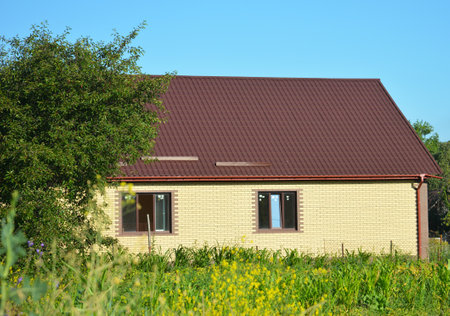 A picturesque view of a small brick house of simple construction with metal dark red roof, rain gutter system with a downpipe, surrounded by trees, green grass and flowers.の写真素材