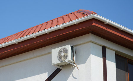 An air conditioner is installed on the house with a metal red roof, soffit and a plastic rain gutter