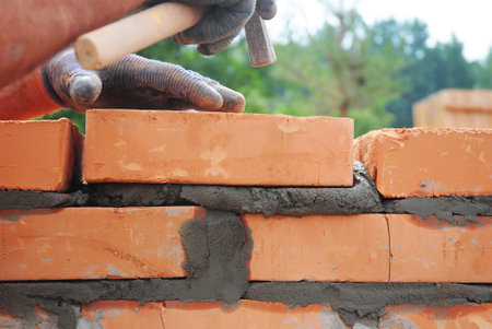 Bricklayer lays bricks on cement mortar. Builder hold brick with hand and knocks with a hammer from above during house wall construction. Bricklaying background with copy space.の写真素材