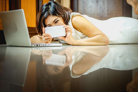 Asia woman using smart phone and laptop lying on the floor at homeの写真素材