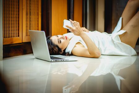 Asia woman using smart phone and laptop lying on the floor at homeの写真素材