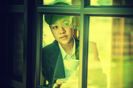 portrait of asia young businessman looking out window while standing in office and drinking coffee...
