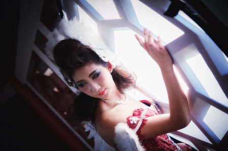 beautiful young woman with white angle wings of red dress posing near windowの写真素材