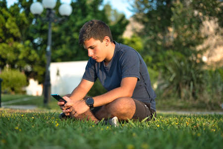 Teenage Boy Using Phone In Urban Setting, Sitting on Grass at sunsetの写真素材