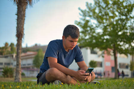 Teenage Boy Using Phone In Urban Setting, Sitting on Grass at sunsetの写真素材
