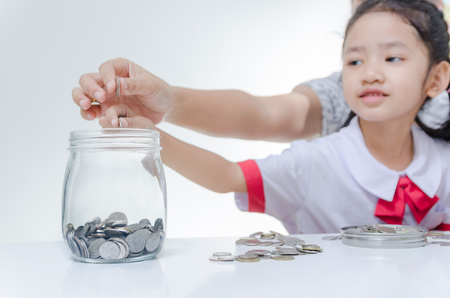 Asian little girl in Thai student uniform putting coin to glass jar with mother hand to saving money focus on hands shallow depth of fieldの写真素材