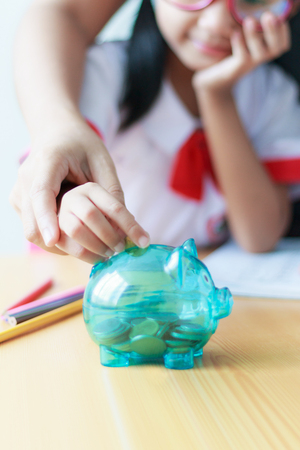 Close up shot Asian little girl in Thai student kindergarten uniform with mother hand putting money coin into clear piggy bank saving money for education concept shallow depth of field select focus on pigの写真素材