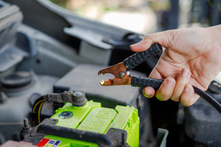 A hand holding a jumper cable clamp close to a car battery, illustrating vehicle maintenance,...