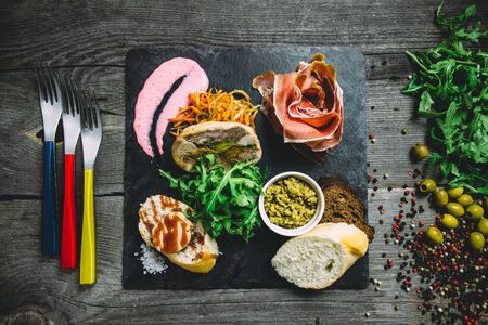 Jamon, meatloaf with mushrooms, bread, arugula, sauce, olives and carrots on a black square plate with forks. Wooden gray background, top viewの写真素材