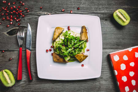 Kiwi carpaccio with tofu cheese, lettuce, arugula, cucumber and pomegranate grains, with herbs and cutlery on square white plate and a gray wooden background, top viewの写真素材