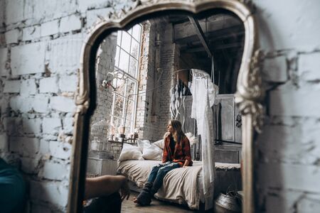 Reflection of the girl in the mirror, she sits on the bed and looking out the window. Reflection in the mirrorの写真素材