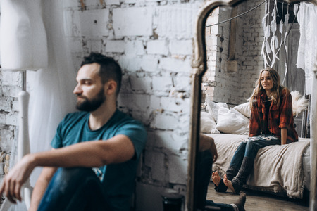 Reflection of a woman in the mirror. Wife sits on the bed in the bedroom and looks at husband, who...