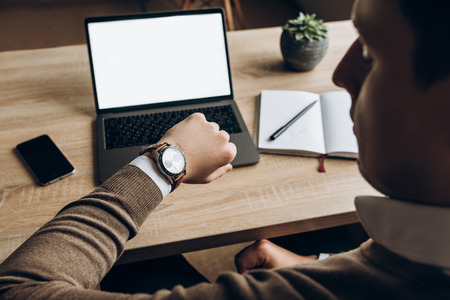 The guy-manager sits at his workplace and looks at the time on his wristwatch. On the desk is a laptop with a clean screen and space for text. Top viewの写真素材