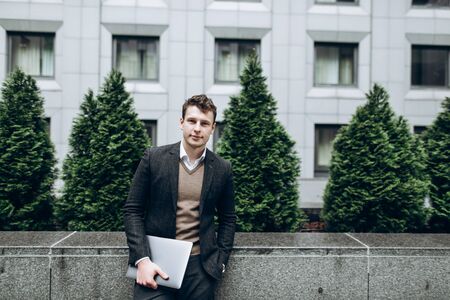 Portrait of a young handsome man in a stylish suit who stands near a business center outdoor with a laptopの写真素材