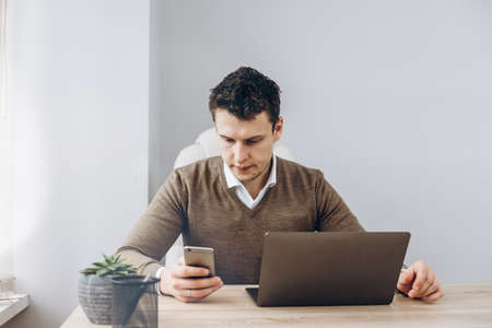 Young businessman works on laptop in his office, and looking for some information in his smartphone