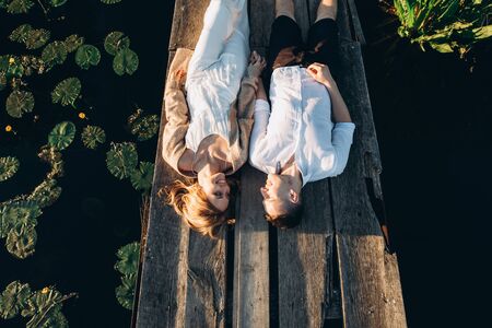 Beautiful young couple lies on a wooden bridge on the lake, looks lovingly at each other and smiles. Top view. Love storyの写真素材